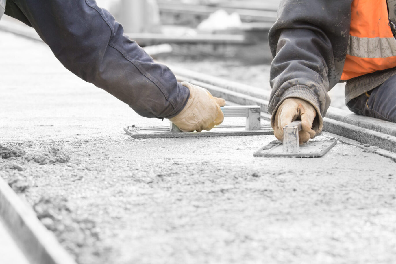 Een cementdekvloer wordt aangebracht door twee vakmannen. De dikke laag cement ligt al, de vakmannen zijn het nauwkeurig glad aan het strijken